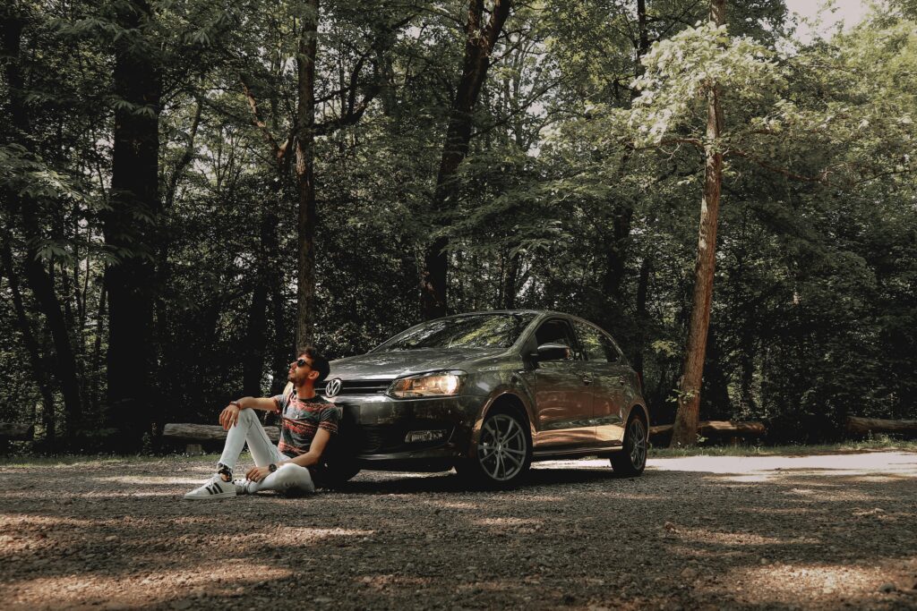 Man in a forest sitting beside his car, enjoying nature and sunlight on a peaceful day.