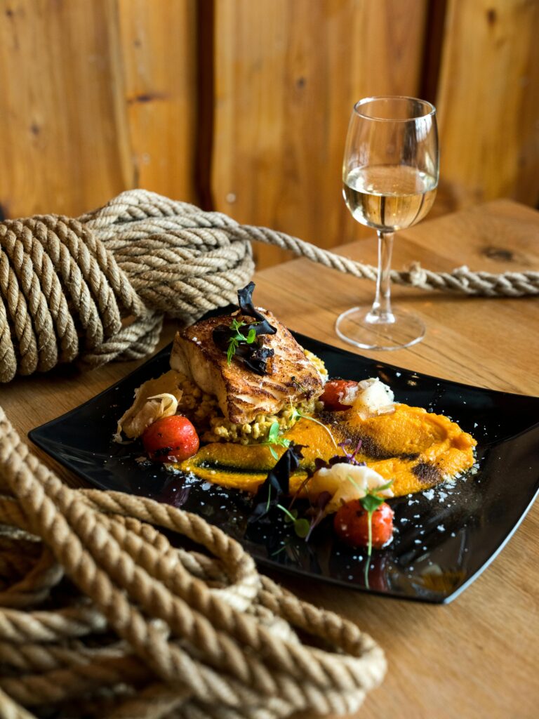 From above of bakery product garnished with orange sauce and strawberries covered with ice cream served on ceramic plate and glass of white wine on wooden table with rope tied into knot
