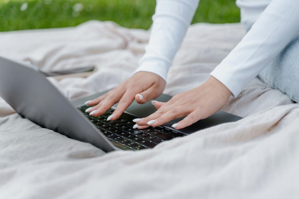 A woman in a white long sleeve shirt typing on a laptop outdoors, enjoying a garden setting.